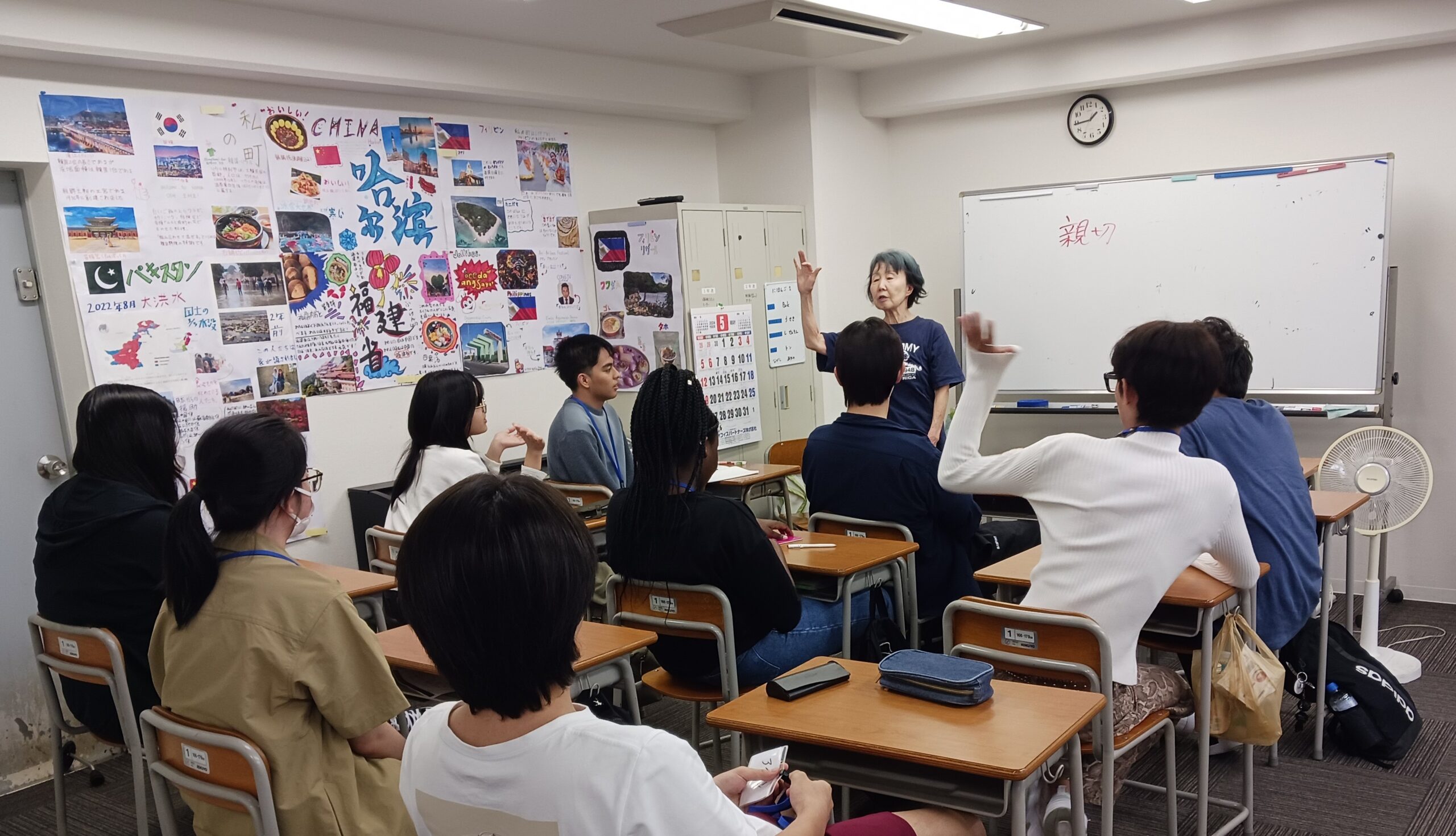 たぶんかフリースクールの授業の様子の写真。ホワイトボードの前に先生が立ち、10人ほどの生徒が席に座って話を聞いている。